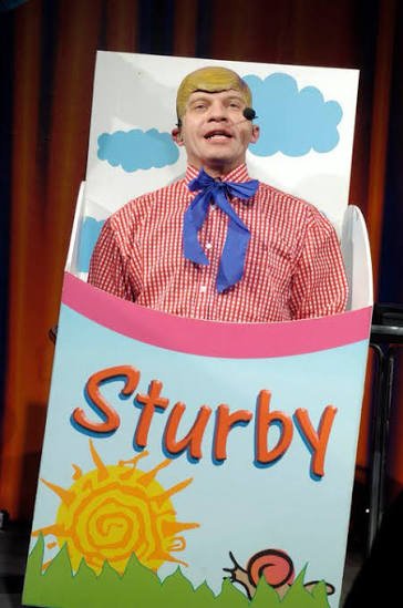 Marco Marzocca nel personaggio di Sturby, con camicia a quadretti rossi e fiocco blu, dietro il cartello con il nome